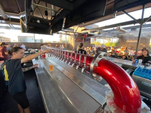 A young person pours a drink from a long, red tap system at a lively outdoor bar with patrons in the background.