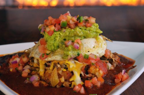 A plate of nachos topped with guacamole, pico de gallo, cheese, and a fried egg, served on a red sauce base.