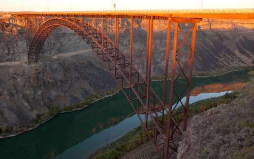 A large metal bridge arches over a river, surrounded by rocky cliffs and colorful foliage at sunset.