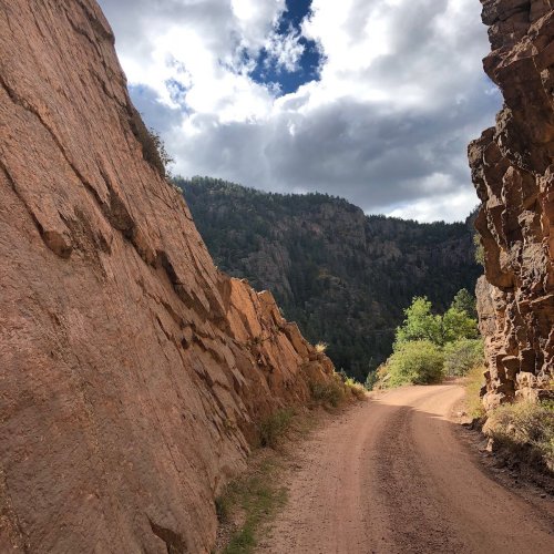 A dirt road winds between rocky cliffs, leading into a lush, green mountainous landscape under a cloudy sky.