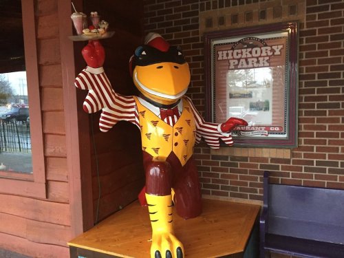A colorful statue of a cartoonish bird in a striped suit, holding a tray with desserts, outside a restaurant.