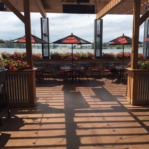 Outdoor dining area with red umbrellas, flower planters, and a view of water under a cloudy sky.