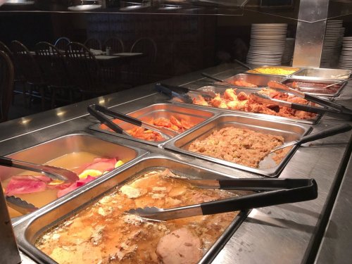 A buffet with various dishes, including meats, sides, and desserts, displayed in metal trays with serving tongs.