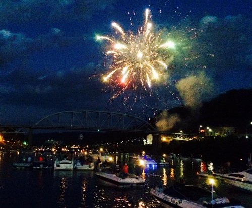 Colorful fireworks burst over a river, illuminating boats and a bridge against a twilight sky.
