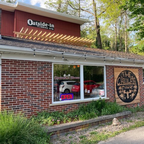 A climbing gym with a sign reading "Outside-In" and a shop called "The Chocolate Moose" nearby, surrounded by greenery.