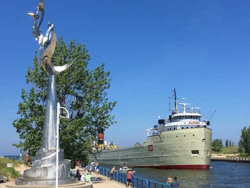 A large ship named "Alpena" sails past a silver sculpture along a waterfront, with people enjoying the sunny day.