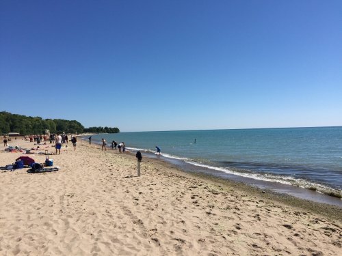 A sunny beach scene with people walking along the shore, sand, and calm water under a clear blue sky.