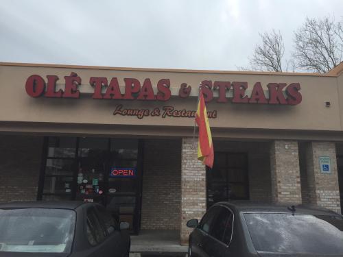 Sign for "Olé Tapas & Steaks Lounge & Restaurant" with an open flag, in front of a building on a cloudy day.