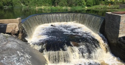 A serene waterfall cascades over a stone dam into a calm river, surrounded by lush greenery.