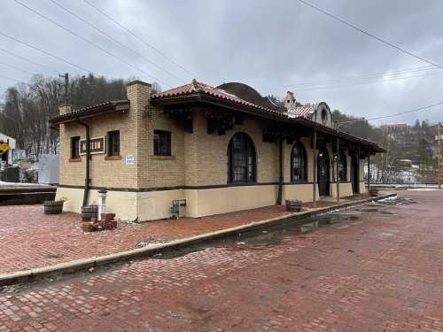 Historic brick train station building with a tiled roof, surrounded by a wet cobblestone area and overcast sky.