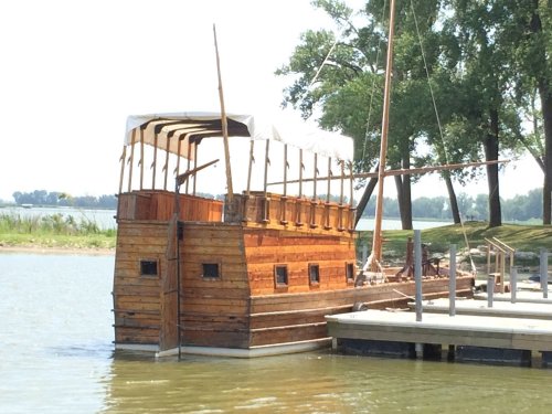 A wooden boat with a canopy docked by a river, surrounded by trees and a calm waterway.