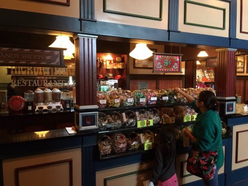 A woman and child browse a colorful candy shop filled with jars and treats behind a wooden counter.