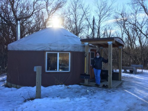 A person stands outside a yurt on a snowy path, surrounded by bare trees and a clear blue sky.