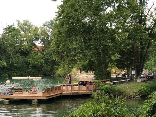 A serene lakeside scene with a wooden dock, people relaxing, and lush green trees surrounding the water.