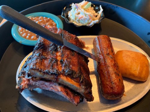 A platter featuring smoked ribs, a sausage link, a roll, baked beans, and coleslaw on a metal tray.