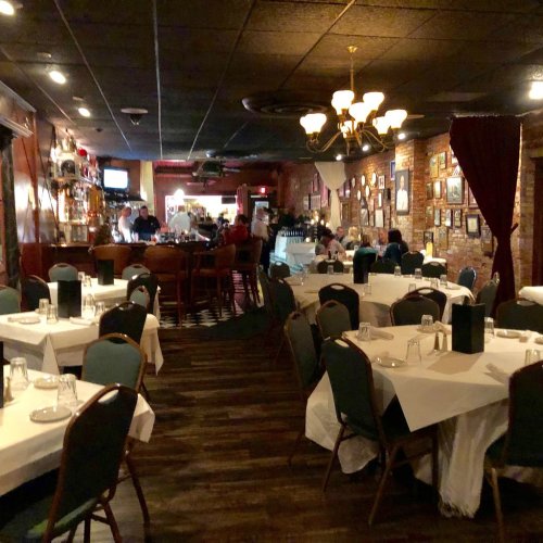 Cozy restaurant interior with tables set for dining, warm lighting, and a bar area in the background.