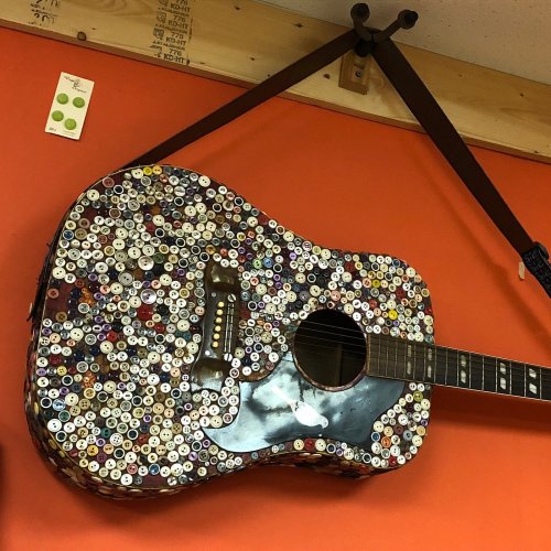 A colorful guitar covered in various buttons, hanging on an orange wall.