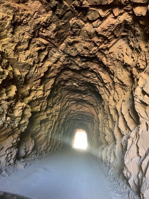 A rocky tunnel with textured walls, leading to a bright light at the end, creating a dramatic perspective.
