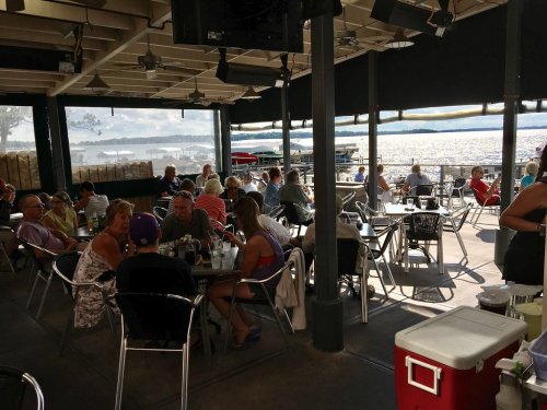 A lively outdoor restaurant scene with diners enjoying meals by the water, surrounded by scenic views.