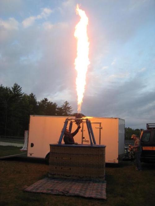 A hot air balloon is being prepared, with a flame shooting upward from the burner against a cloudy sky.