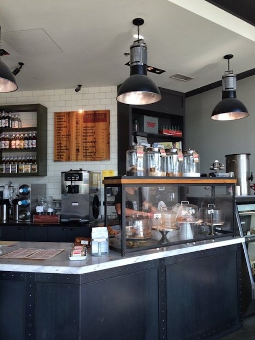 A modern café interior featuring a counter with pastries, coffee equipment, and shelves of drinks in a bright setting.