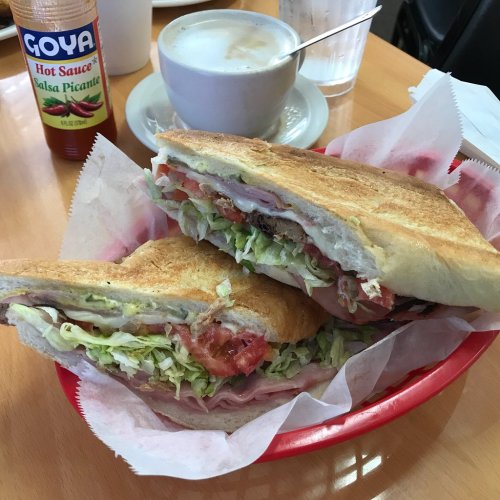 A sandwich filled with lettuce, tomatoes, and ham, served in a basket alongside a cup of coffee and hot sauce.