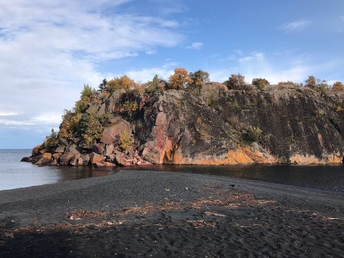A rocky shoreline with autumn foliage, featuring a black sand beach and calm water under a partly cloudy sky.