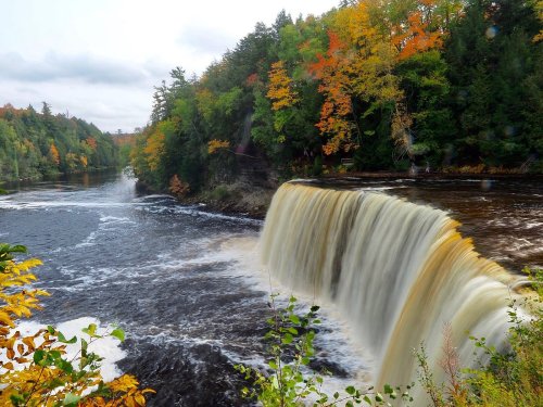 A scenic waterfall surrounded by vibrant autumn foliage and a flowing river, under a cloudy sky.