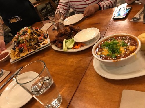 A table with a platter of nachos, chicken wings, a bowl of chili topped with cheese, and a glass of water.