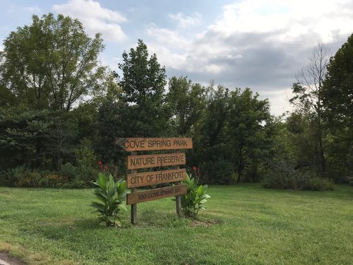 Sign for Cove Spring Park and Nature Preserve in Framingham, surrounded by greenery and trees under a cloudy sky.