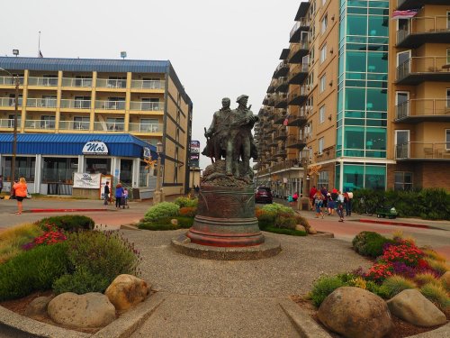 A bronze statue of two soldiers stands in a plaza, surrounded by flowers and buildings, with people walking nearby.