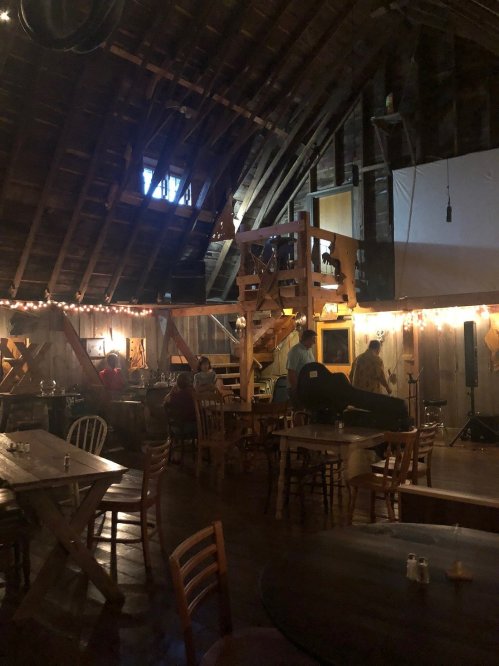 Interior of a rustic barn with wooden beams, tables, and a staircase, softly lit with string lights.