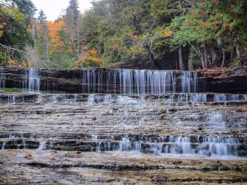 A serene waterfall cascades over rocky steps, surrounded by autumn foliage and lush greenery.