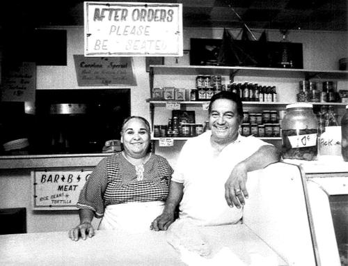 A smiling couple stands behind a counter in a small grocery store, surrounded by shelves of canned goods and signs.