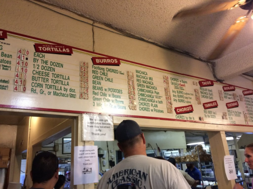 Menu board displaying various tortilla and burrito options in a casual dining setting. Customers are in line.