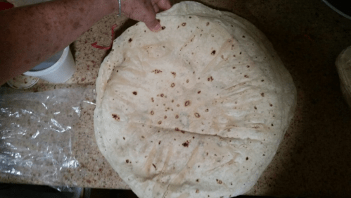 A hand holds a large, round tortilla on a countertop, showcasing its texture and size.