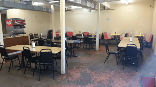 Empty diner with several tables and chairs, a soda fountain in the background, and a worn floor.