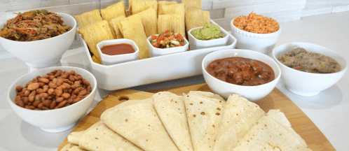 A spread of tamales, tortillas, beans, salsa, guacamole, and shredded meat in bowls on a wooden board.