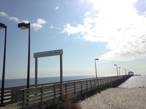 A wooden pier extends into the ocean under a bright sky, with a sign reading "Lighthouse Pier" visible.