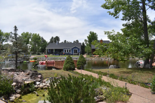 A serene landscape featuring a pond, lush greenery, and a blue house in the background under a partly cloudy sky.