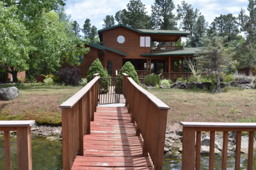 A wooden bridge leads to a two-story cabin surrounded by trees and greenery, with a stream in the foreground.