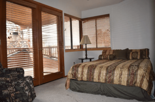 Cozy bedroom with a bed, decorative bedding, a lamp, and large windows with wooden blinds.