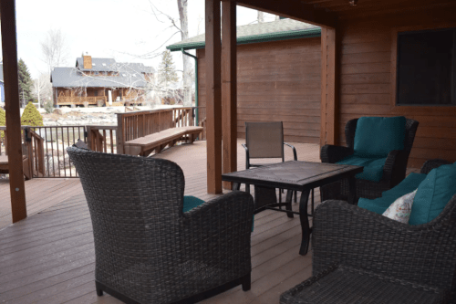 Cozy outdoor seating area with wicker chairs and a table on a wooden deck, surrounded by trees and a distant house.