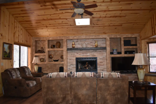 Cozy living room with wooden walls, a stone fireplace, and comfortable seating. Natural light from windows.