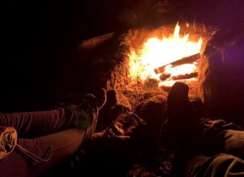 Two pairs of feet resting near a glowing campfire, surrounded by darkness. Flames illuminate the scene.