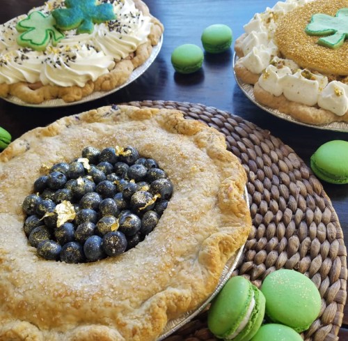 Three decorative pies with toppings, surrounded by green macarons, on a woven placemat.