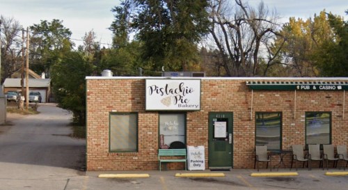 A brick building with a sign reading "Pistachio Pie Bakery" and outdoor seating, surrounded by trees and a parking area.