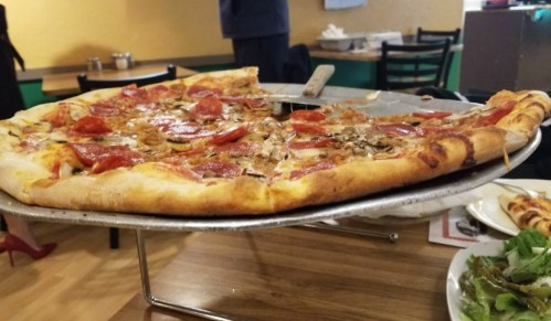 A large pizza topped with pepperoni, vegetables, and cheese, served on a metal stand in a casual dining setting.