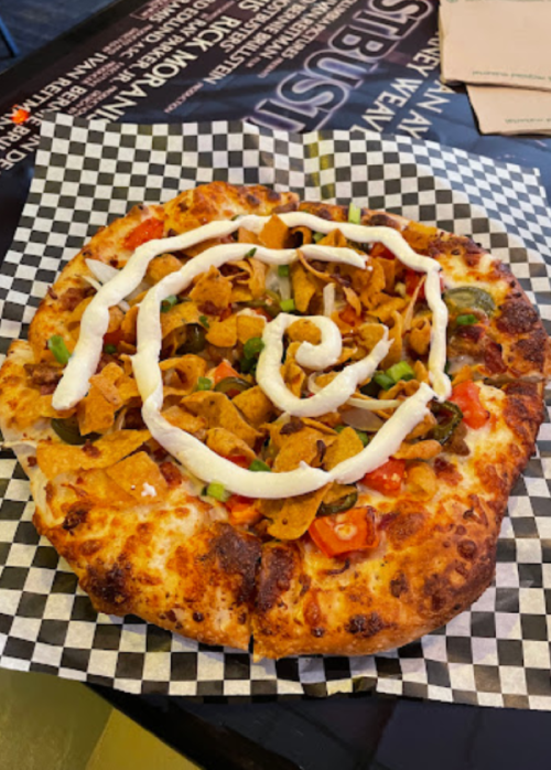 A pizza topped with cheese, tomatoes, green peppers, Fritos, and a swirl of sour cream on a checkered paper.