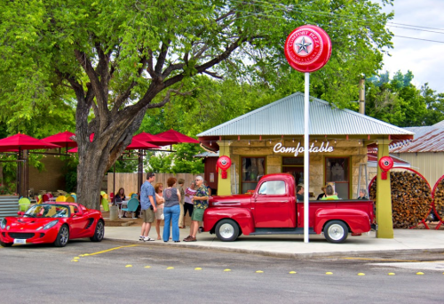 A red vintage truck parked outside a cozy eatery with outdoor seating and a large tree, surrounded by people.